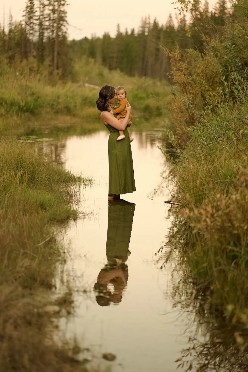 Mother holding her baby beside a still pond that mirrors them both