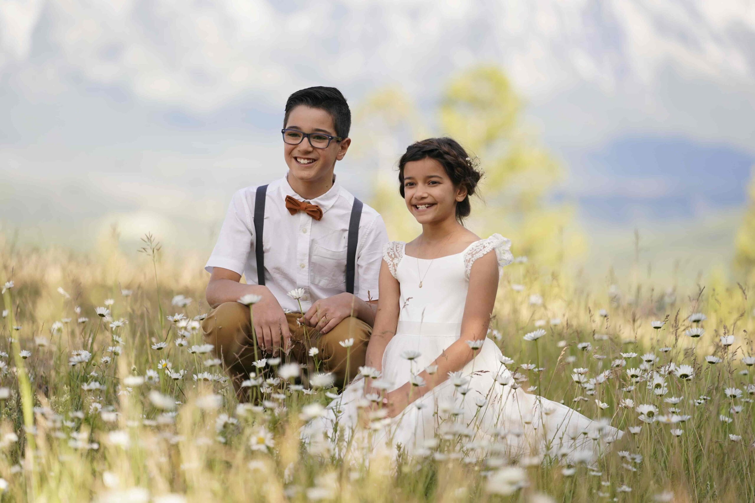 Two siblings dressed up sitting together in a daisy-filled meadow with mountains in the background