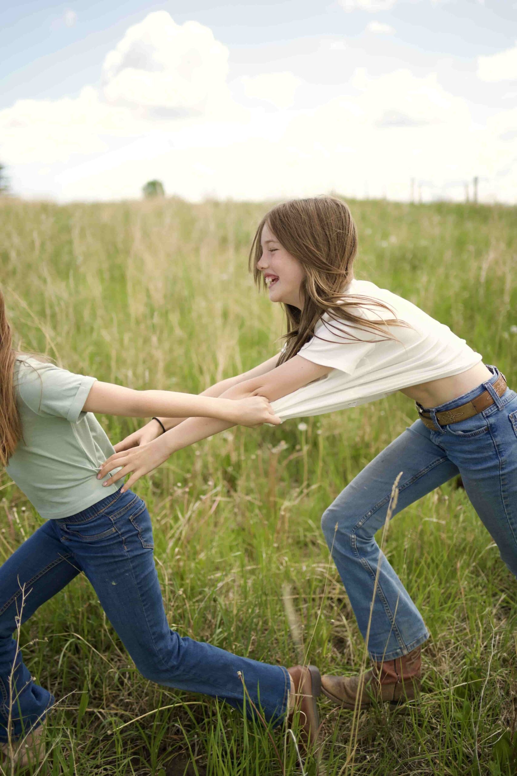 Two sisters laughing and playing tug-of-war with hands in a grassy field